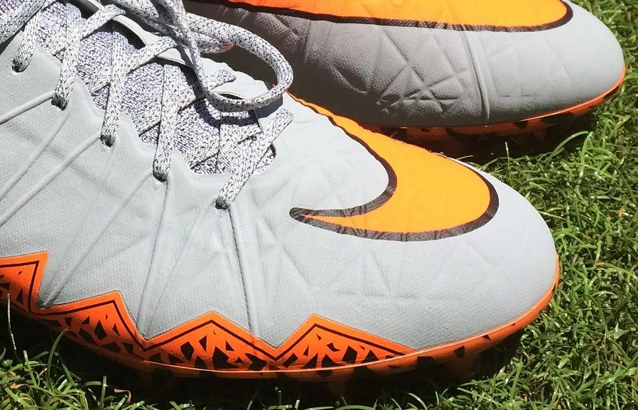 Close-up of gray and orange Nike football cleats on grass, showing a wide forefoot shape, textured upper, and molded studs for traction.