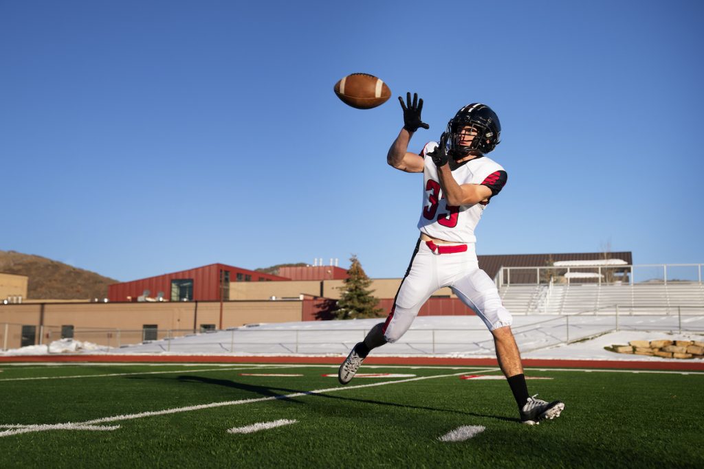 American football player in white uniform jumping to catch a pass on a turf field, wearing black cleats during game action under clear blue sky.